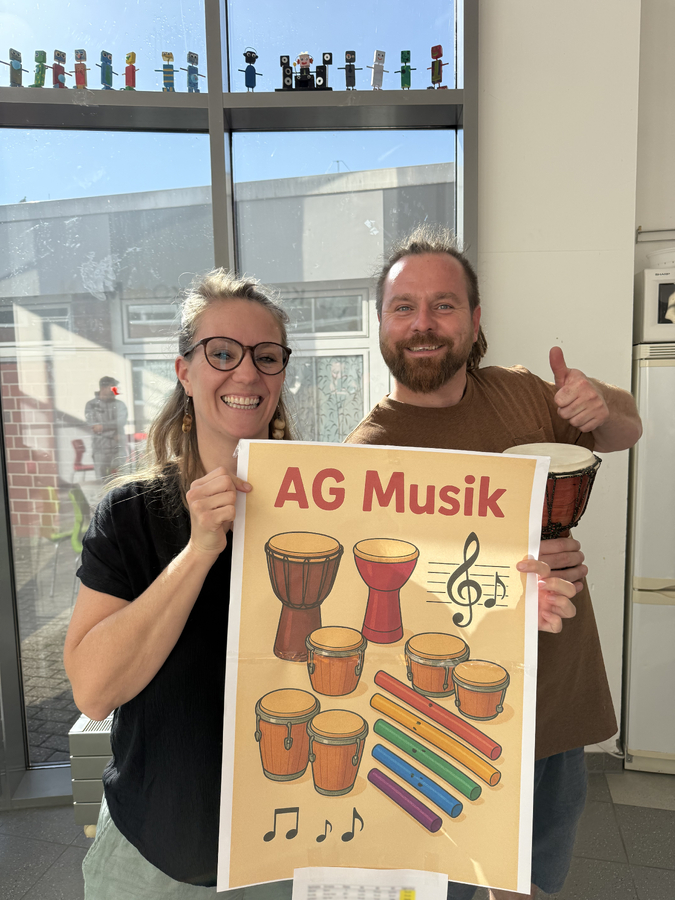 Eine Lehrerin und ein Lehrer halten ein Plakat mit der Schrift "AG Musik" hoch. Auf dem Plakat sind verschiedene Trommeln, Klangrohre und Noten zu sehen.